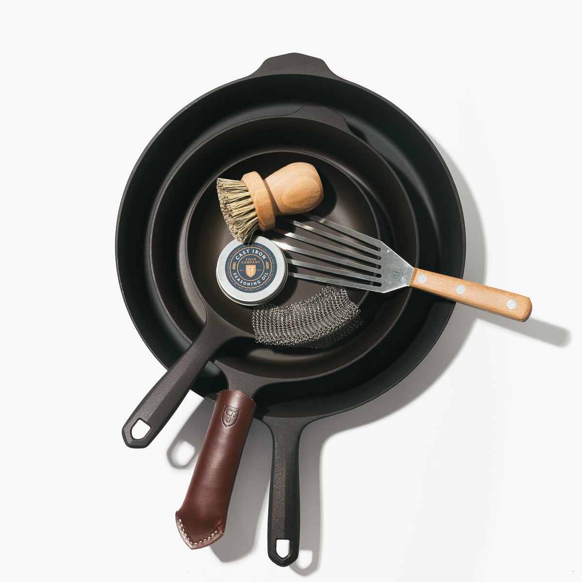 Three cast iron skillets showing a slotted turner, wooden brush, seasoning oil, mesh scrubber, and leather handle cover inside.