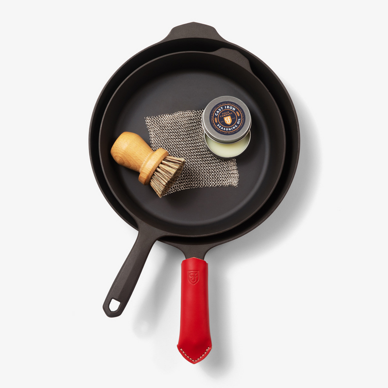 Nested Field Skillets with scrub brush, chain mail, seasoning oil tin, and a red leather handle cover, overhead view