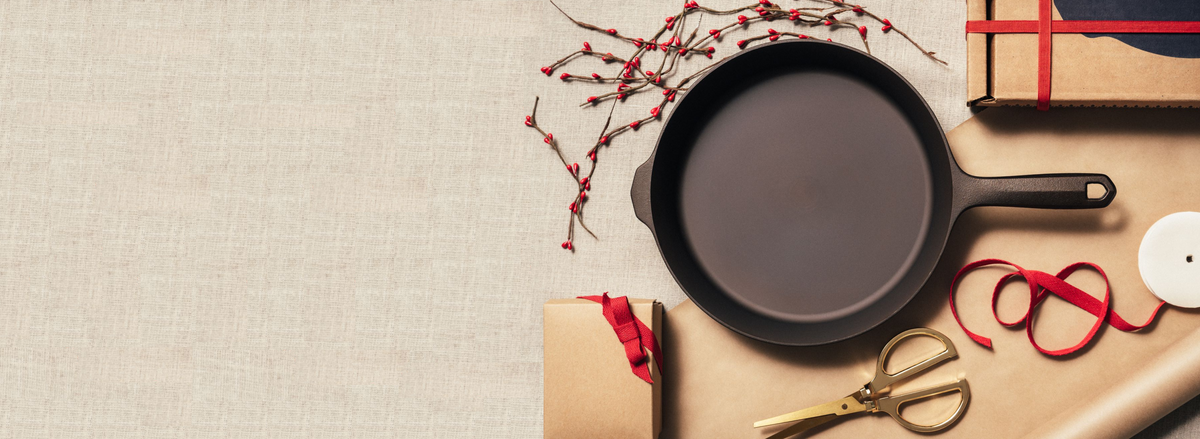 Overhead of a Field Skillet on linen with red berry sprigs, wrapped gifts, ribbon, and brass scissors